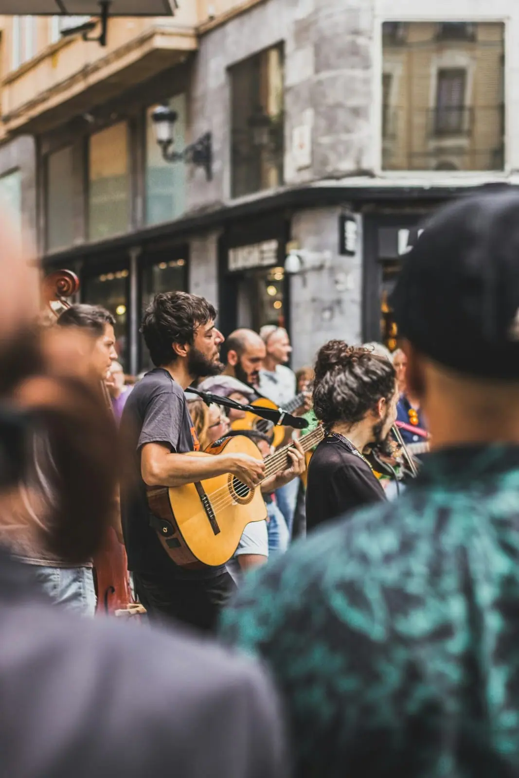 Músicos tocan la guitarra y el violín en una calle comercial, rodeados de vecinos que se paran a escucharlos