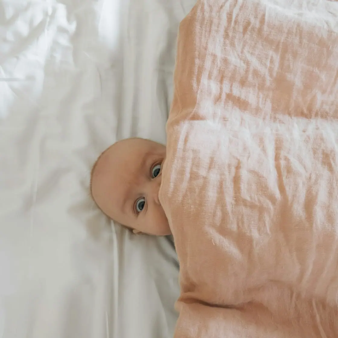 Un bebé tumbado sobre una sábana blanca asoma la cabeza bajo una manta rosa.