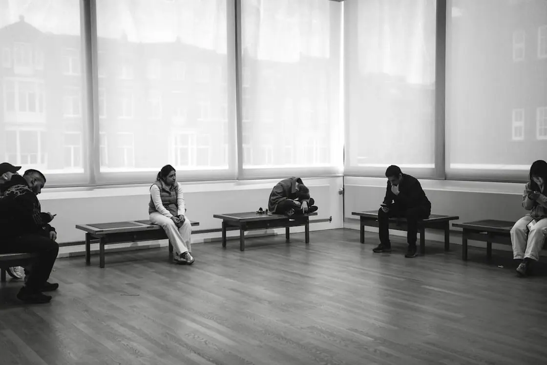 Sala de espera en blanco y negro con varias personas sentadas en bancos, cada una por su lado.