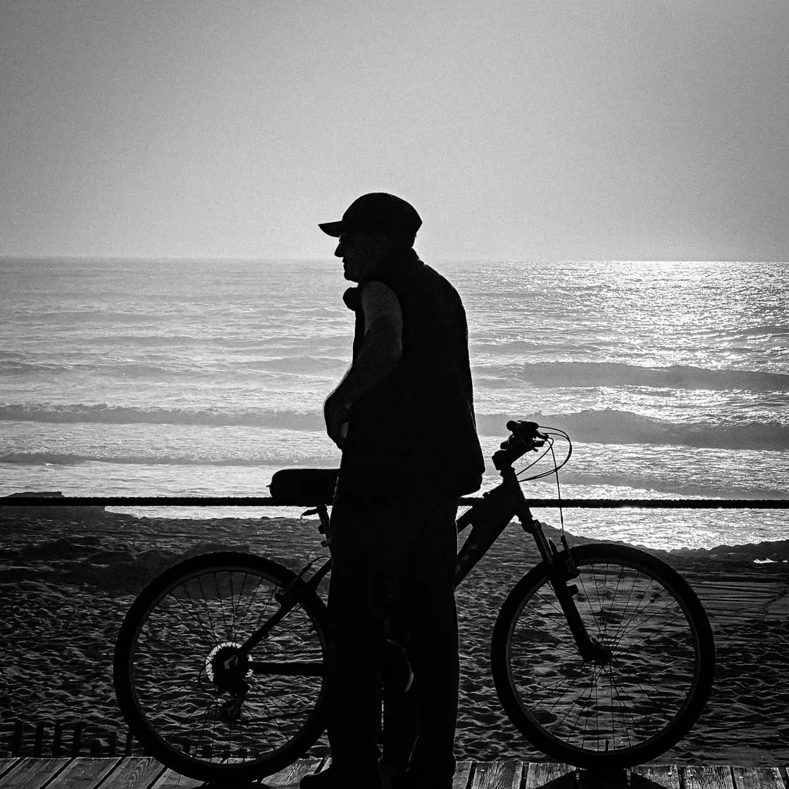 Foto en blanco y negro de un hombre mayor con gorra, a contraluz, junto a su bicicleta en un paseo frente al mar.