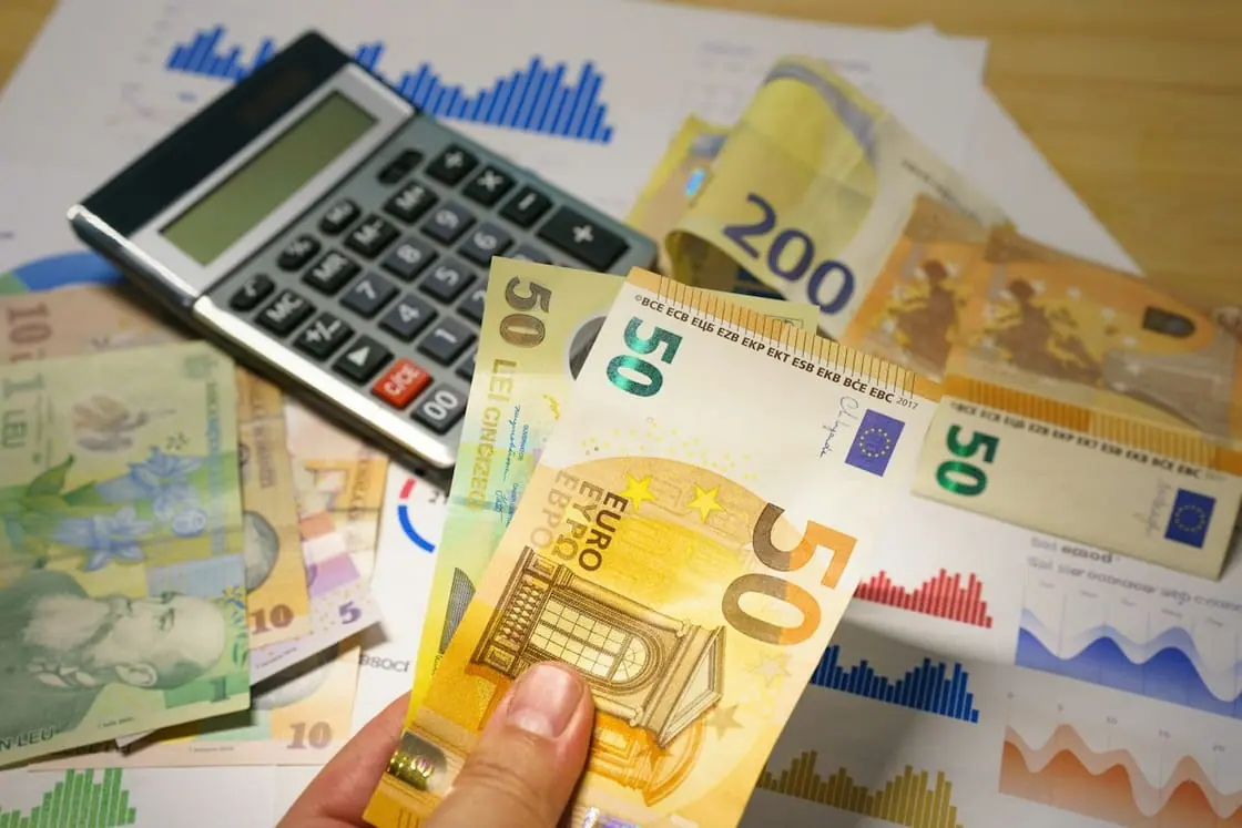 Una mano sostiene billetes de 50 euros junto a una calculadora y unos gráficos impresos.
