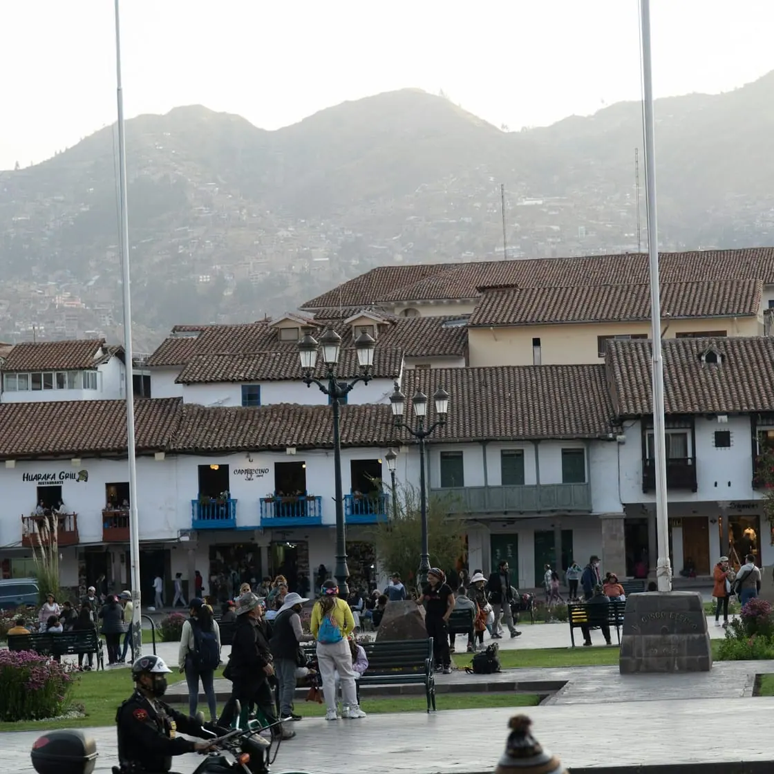 Una plaza con soportales y montañas al fondo; en primer plano, un hombre en moto y gente paseando.