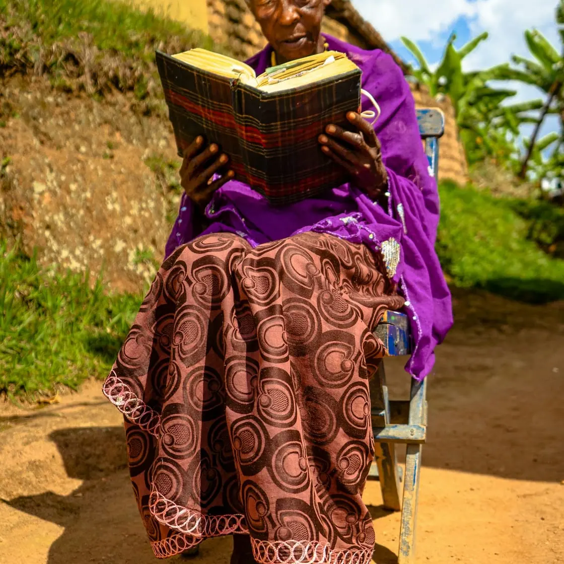Una mujer mayor sentada en una silla al aire libre, en un camino de tierra, lee un libro.