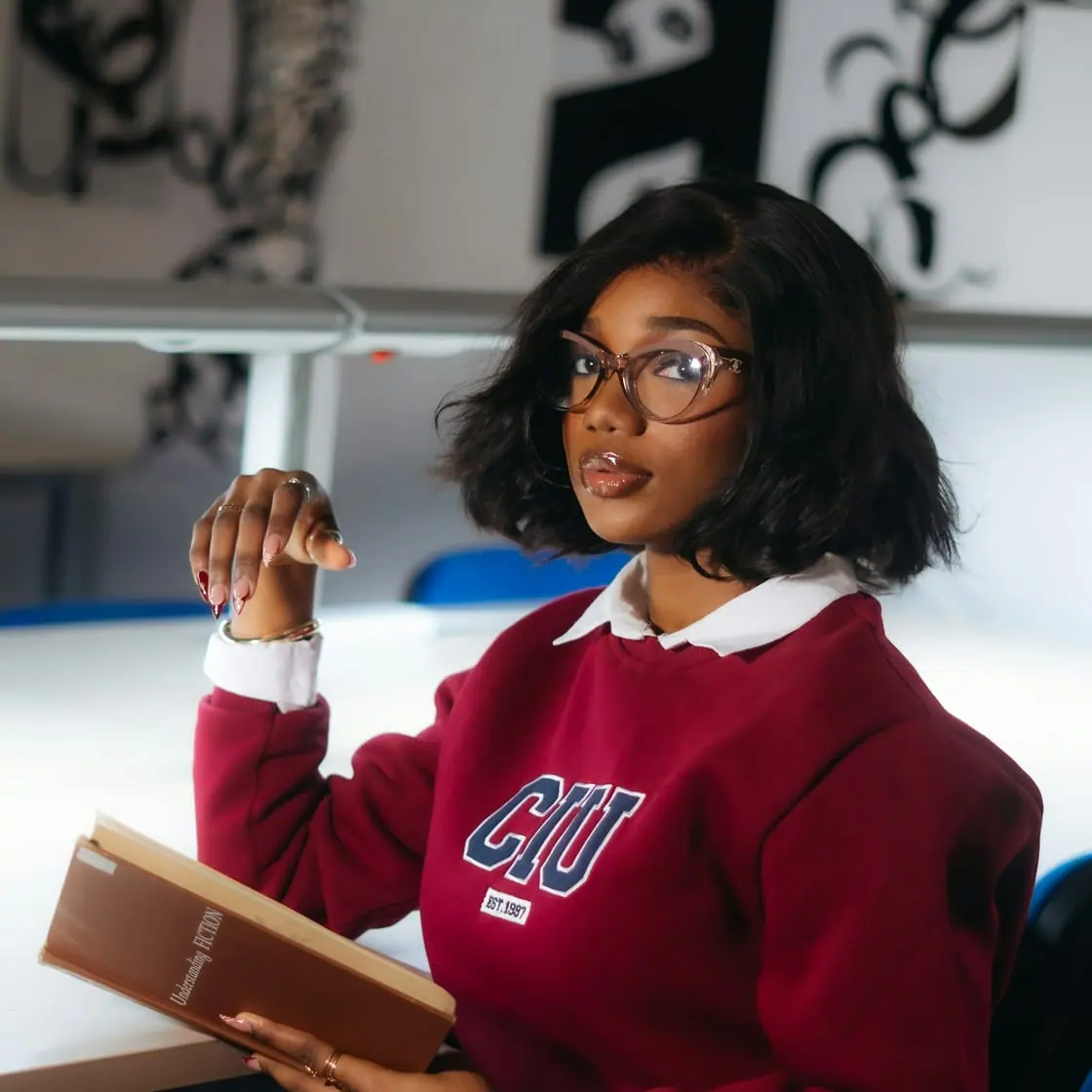 Una estudiante con gafas y sudadera granate sostiene un libro abierto mientras atiende en clase.