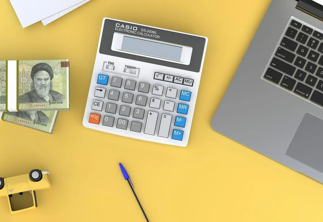 Una calculadora, unos billetes, un bolígrafo y un portátil sobre una mesa amarilla, vistos desde arriba.