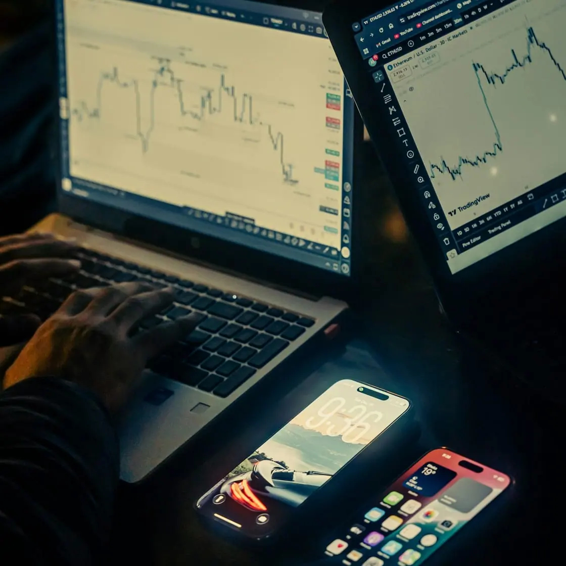 Dos portátiles con gráficos de bolsa en la pantalla y dos móviles encendidos sobre una mesa a oscuras.