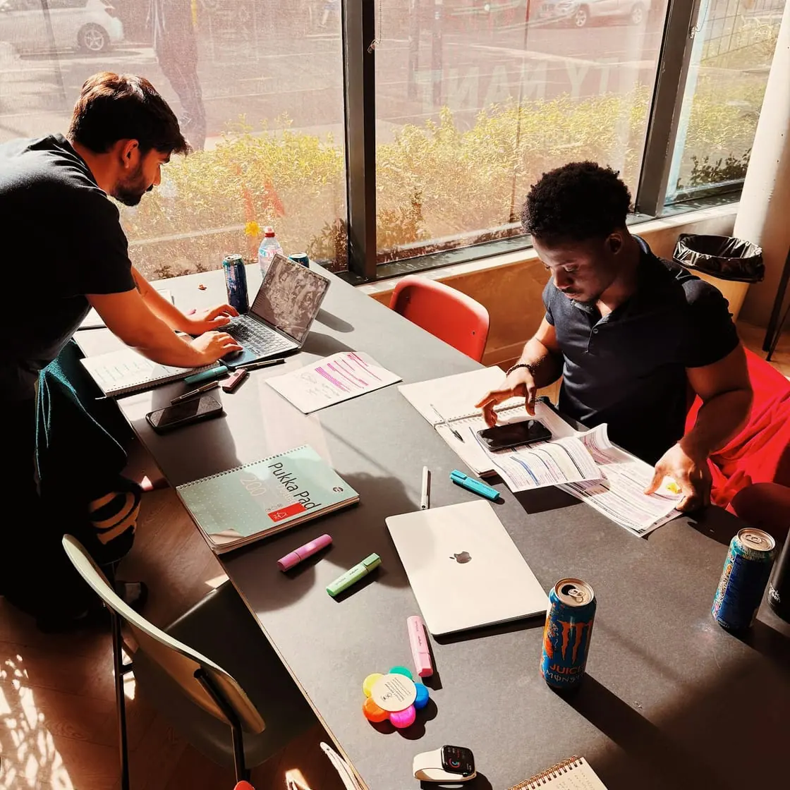 Varios estudiantes trabajan alrededor de una mesa llena de apuntes, portátiles y rotuladores junto a un ventanal.