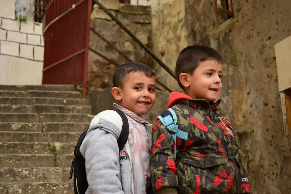Dos niños pequeños con mochila sonríen al pie de unas escaleras de piedra.