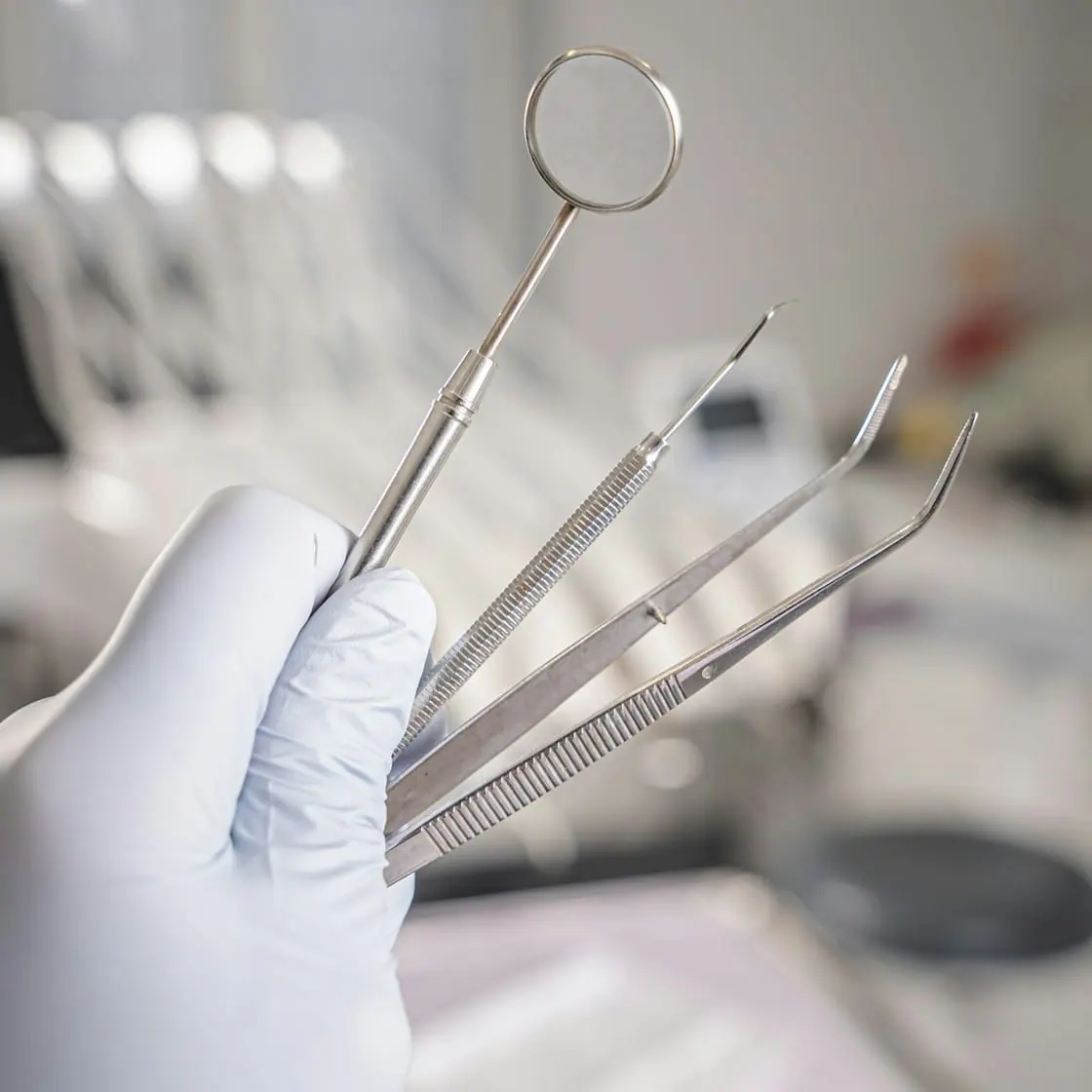 Una mano con guante sostiene instrumentos de dentista, con el sillón de la consulta al fondo.