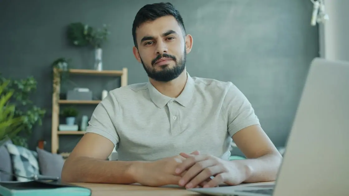 Un hombre con barba y polo claro, sentado a una mesa delante de un portátil, mira a la cámara con las manos juntas.