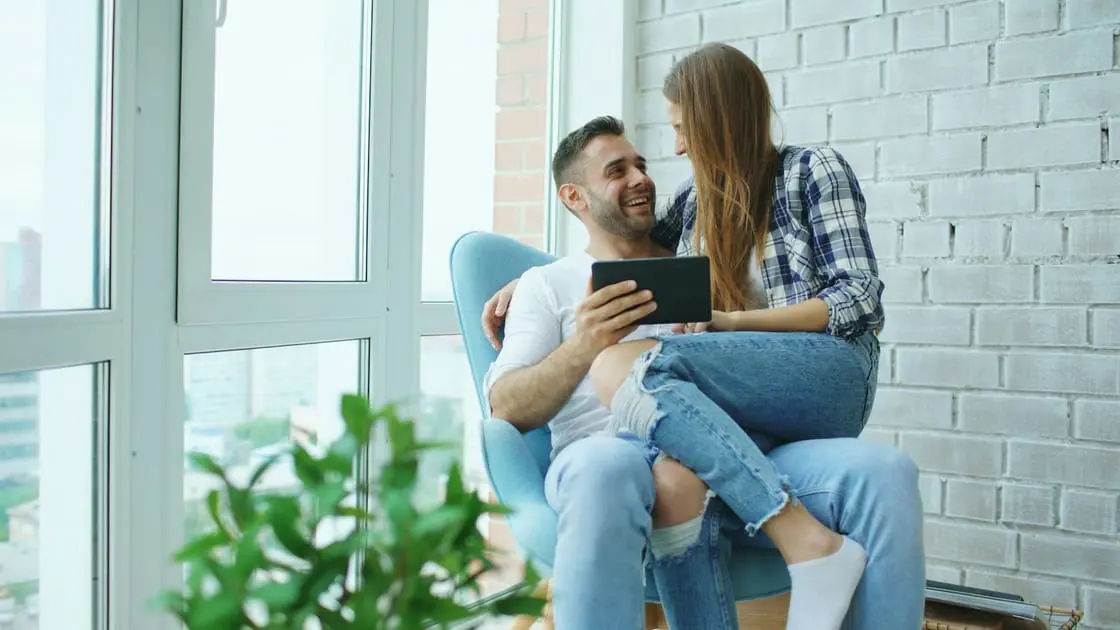 Una pareja joven sonríe sentada en un sillón junto a un ventanal mientras mira una tableta.