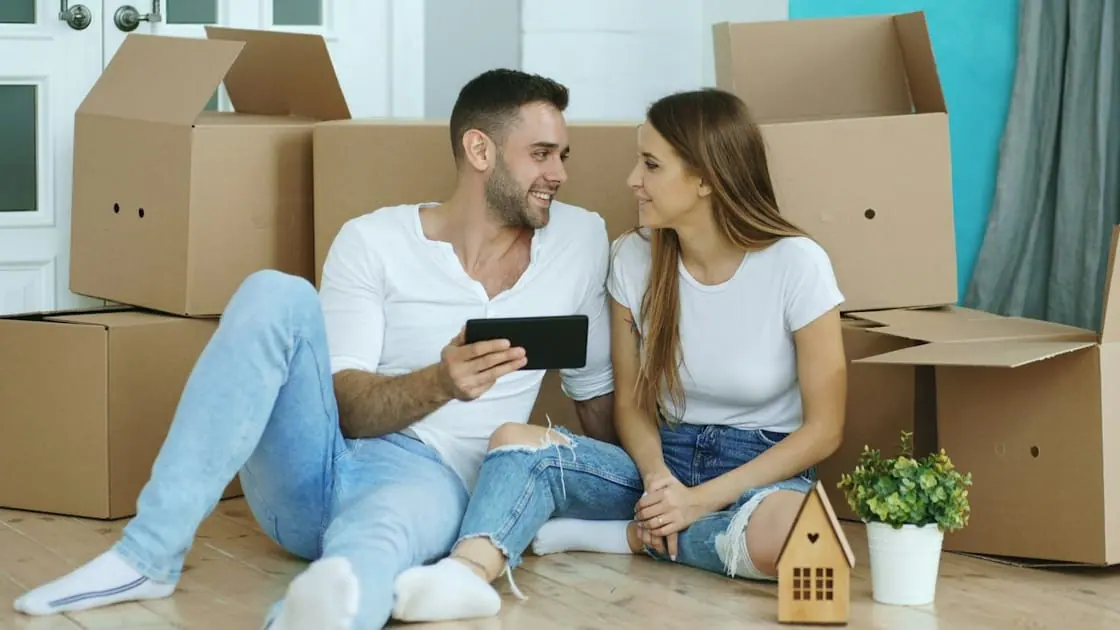 Una pareja joven sentada en el suelo entre cajas de mudanza mira una tableta, con una casita de madera a su lado.
