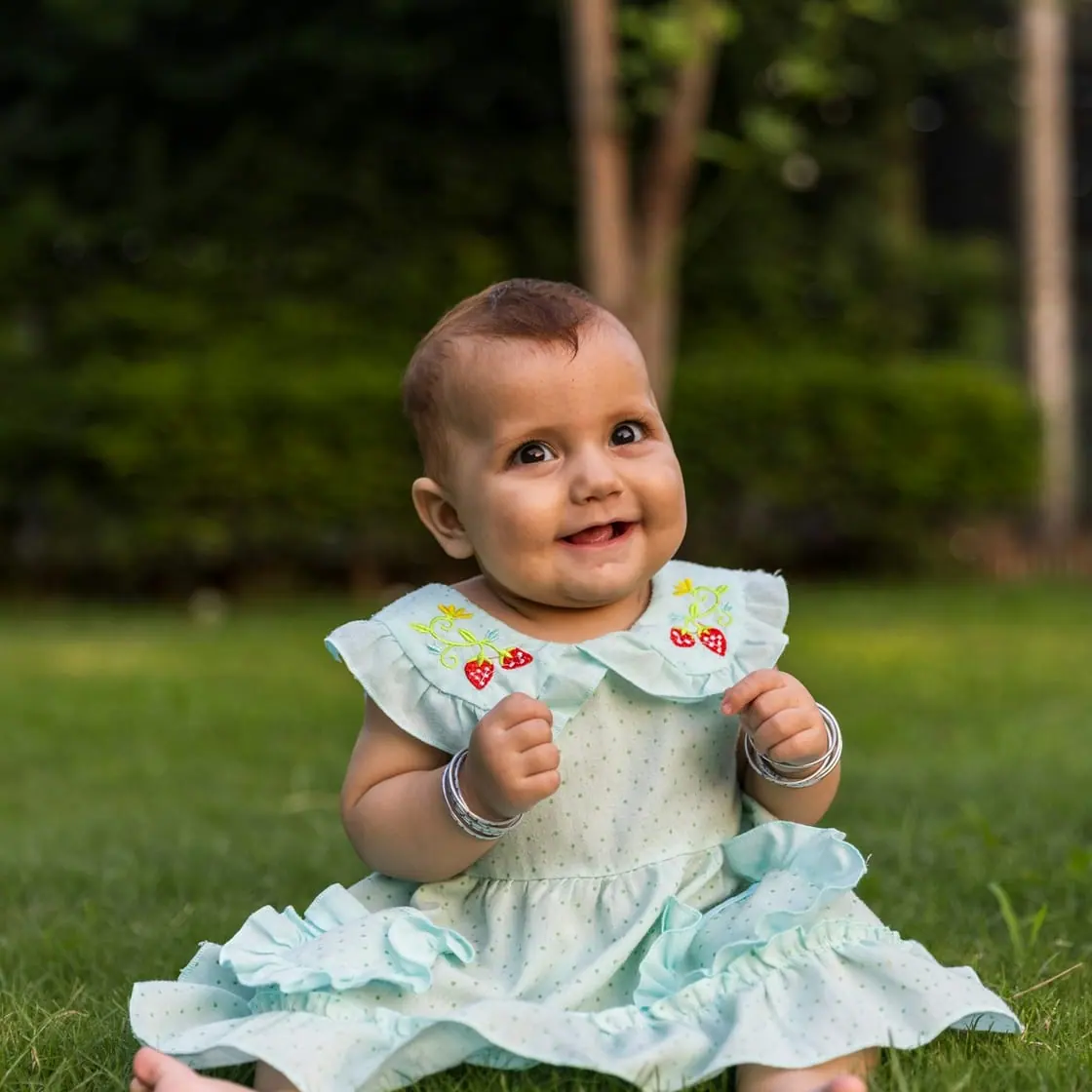 Una bebé sonriente con vestido claro, sentada descalza sobre el césped de un jardín.