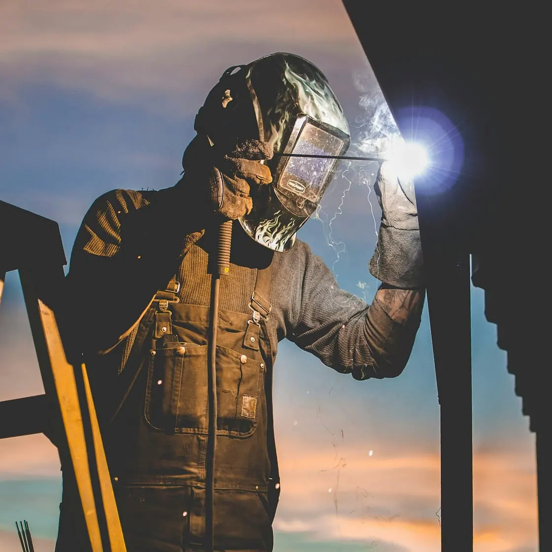 Un soldador con careta protectora trabaja en lo alto de una estructura metálica al atardecer.