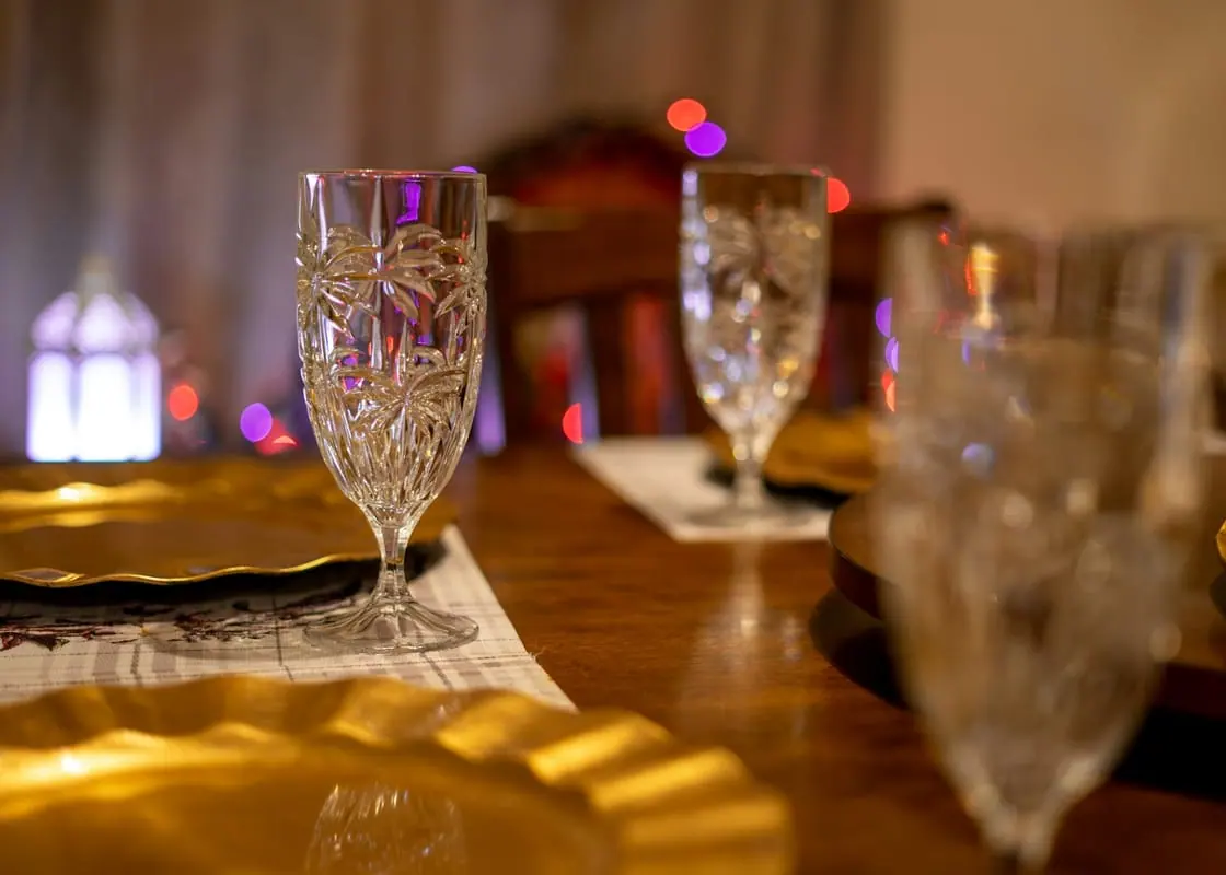 Copas de cristal tallado y platos dorados sobre una mesa puesta, con luz cálida de fondo.