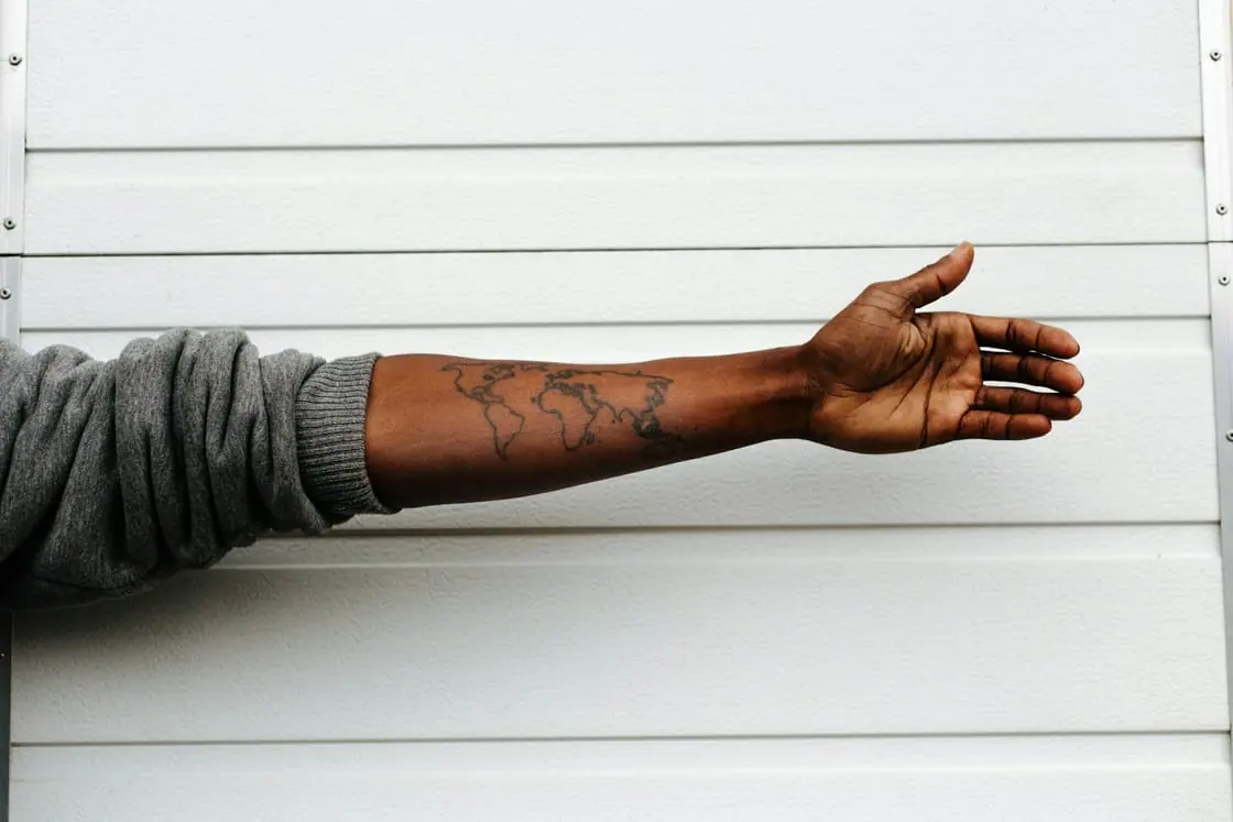 Un brazo tatuado con un mapamundi se extiende con la palma abierta delante de una pared blanca de lamas.