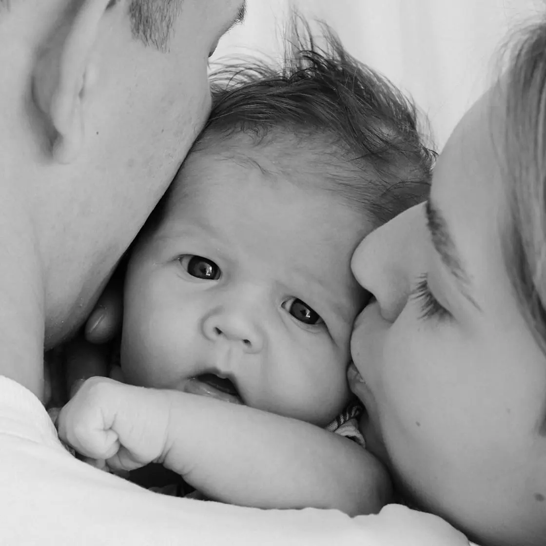 Foto en blanco y negro de un padre y una madre besando a su bebé en las mejillas.