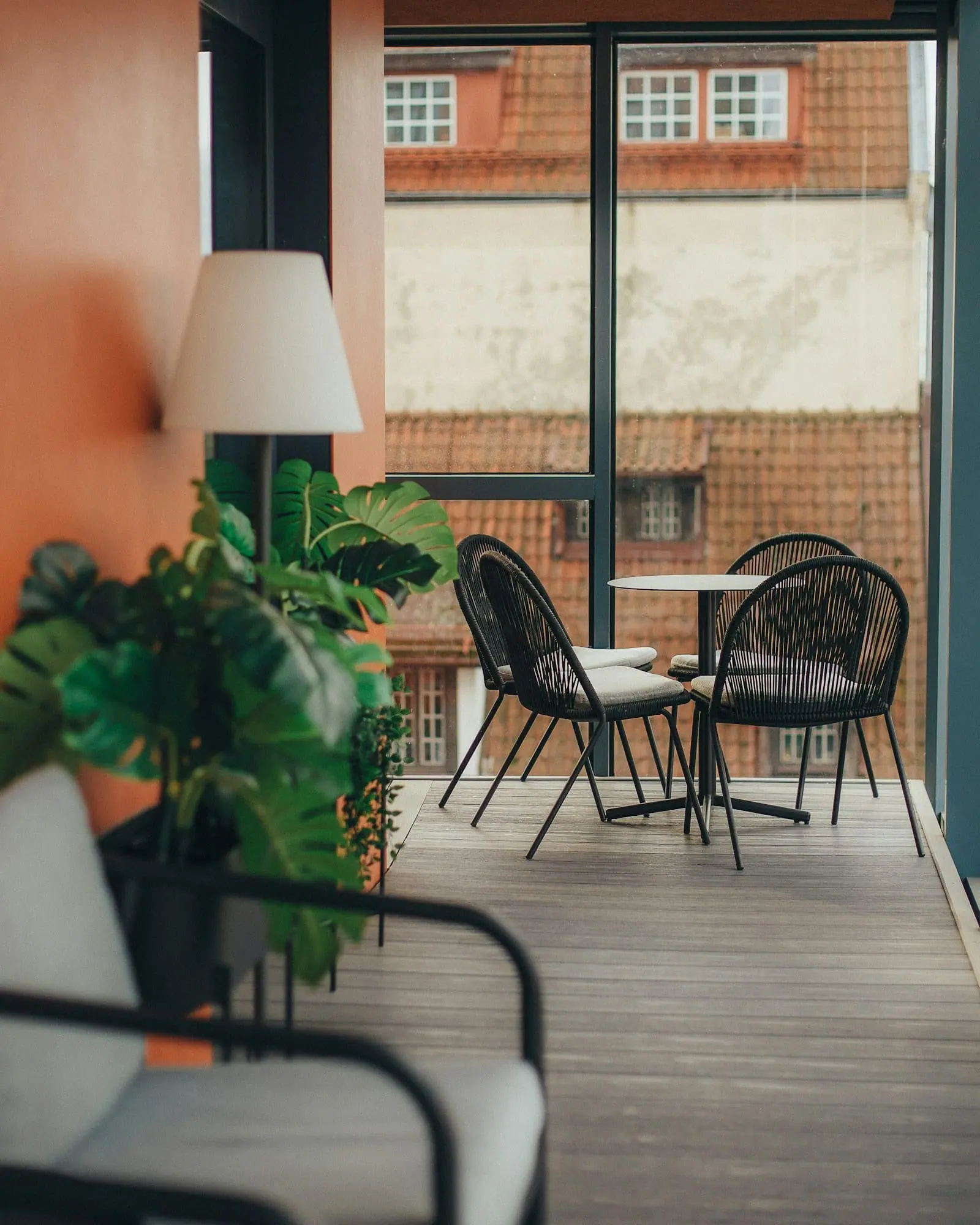 Una terraza acristalada con sillas, una mesa redonda y plantas, con vistas a otros edificios de ladrillo.