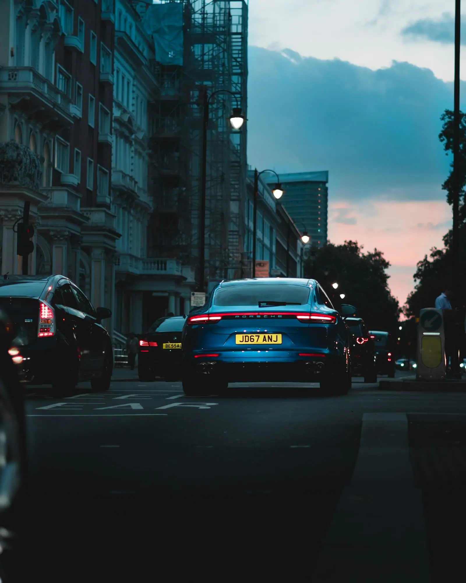 Una calle de ciudad al atardecer, con coches circulando entre edificios antiguos y farolas encendidas.