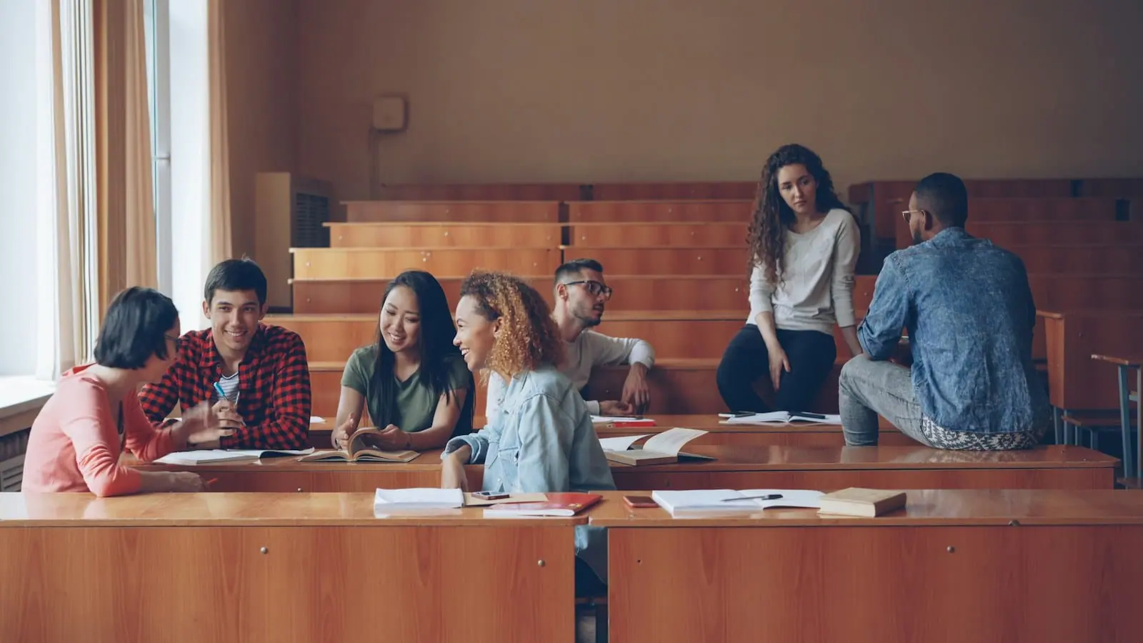 Un grupo de estudiantes charla sentado en los bancos de madera de un aula universitaria en gradas.