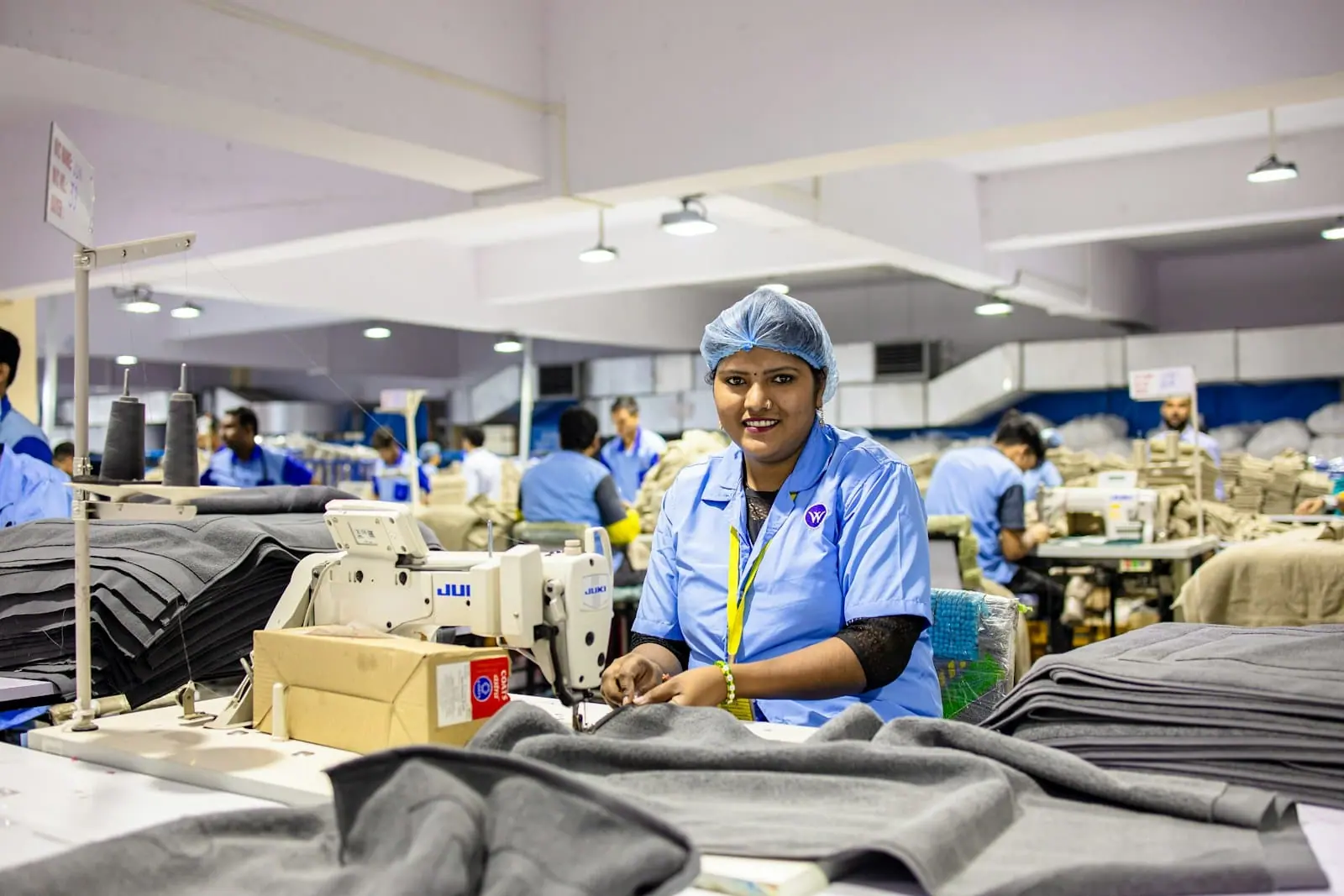 Una trabajadora con bata y gorro azules sonríe junto a su máquina de coser en una gran nave textil llena de compañeros.