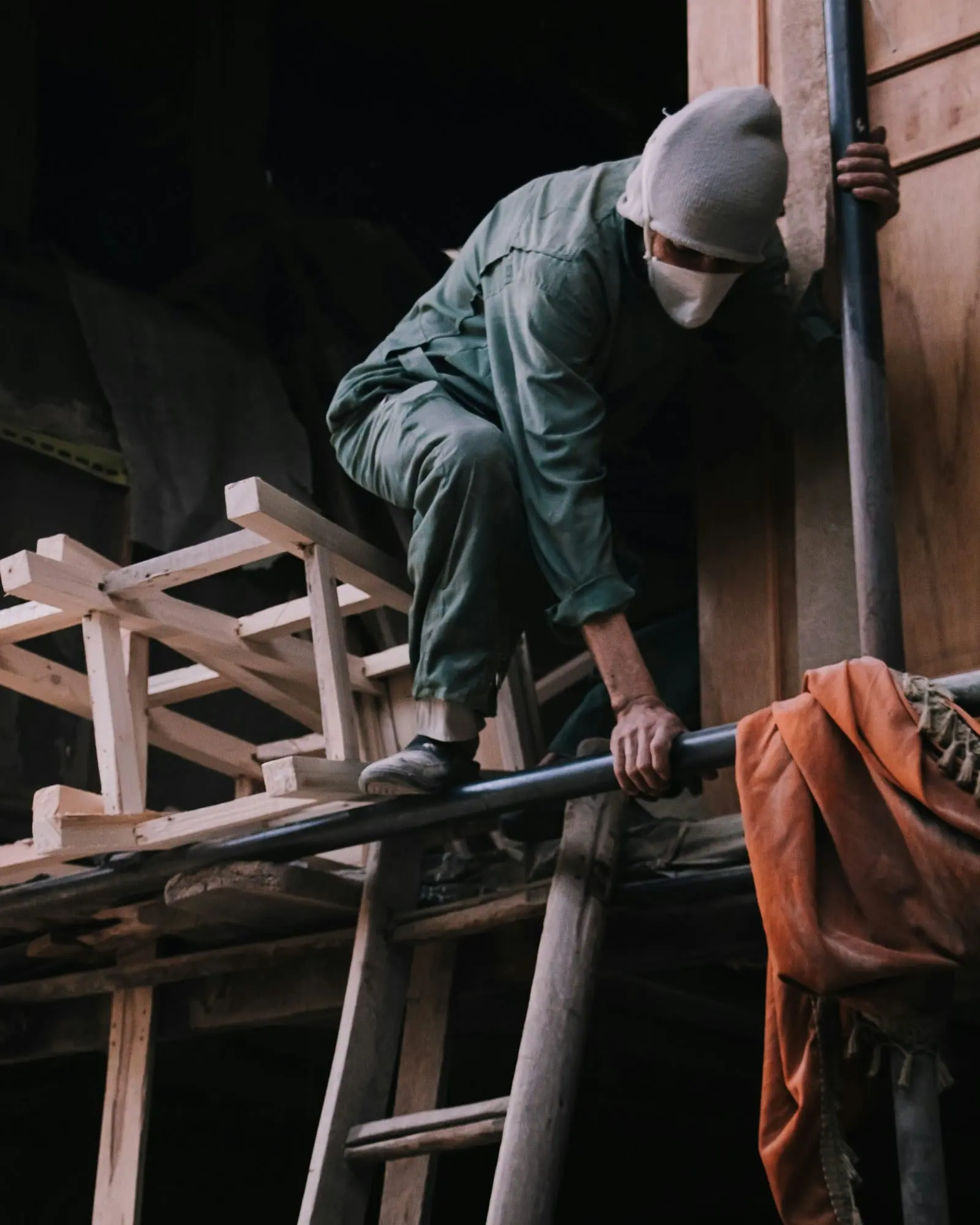 Un trabajador con gorro de lana y mascarilla sube por una escalera de madera en un taller de techo alto.